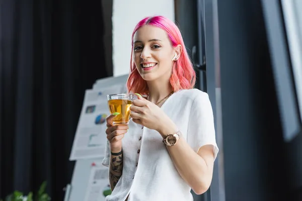 Positive Geschäftsfrau mit rosa Haaren und Kopfhörer hält Tasse Tee im Büro — Stockfoto