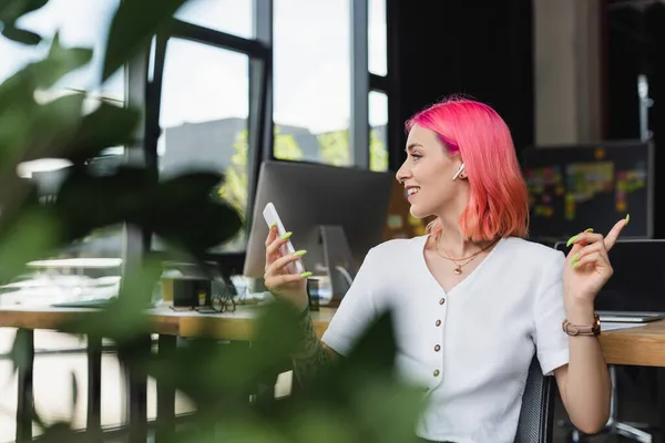 Улыбающаяся молодая деловая женщина с розовыми волосами и наушниками с помощью смартфона в офисе — стоковое фото