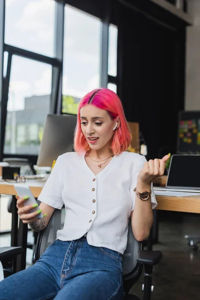 Jeune femme d'affaires tatouée avec des cheveux roses et écouteur en utilisant smartphone dans le bureau — Photo de stock