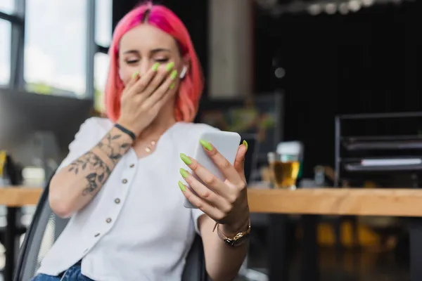 Blurred businesswoman with pink hair and earphone using smartphone and covering mouth while laughing in office — Stock Photo