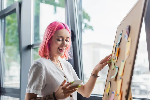 Glückliche Geschäftsfrau mit rosafarbenen Haaren hält Smartphone in der Hand und zeigt mit klebrigen Zetteln auf die Tafel — Stockfoto
