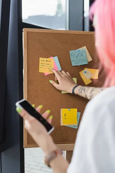 Blurred businesswoman with pink hair holding smartphone with blank screen and pointing at board with sticky notes — Stock Photo