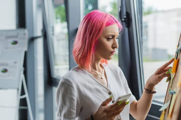 Businesswoman with pink hair holding smartphone and pointing at board with sticky notes — Stock Photo