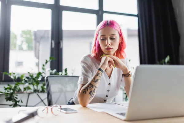 Geschäftsfrau mit pinkfarbenen Haaren blickt auf Smartphone neben Laptop auf Schreibtisch — Stockfoto