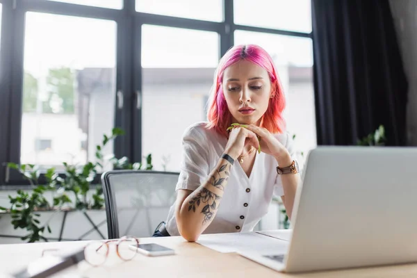 Donna d'affari con i capelli rosa guardando smartphone vicino al computer portatile sulla scrivania — Foto stock