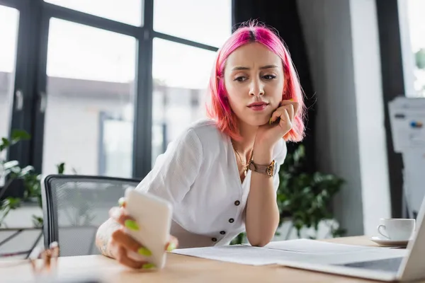 Smiling businesswoman with colorful hair looking at smartphone near laptop — Stock Photo