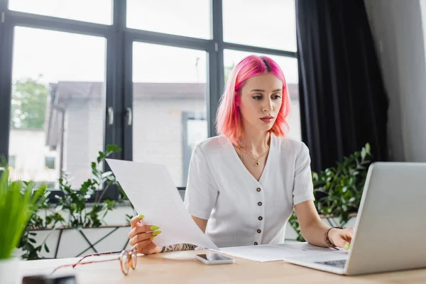 Geschäftige Geschäftsfrau mit buntem Haar hält Dokument in der Hand und benutzt Laptop — Stockfoto
