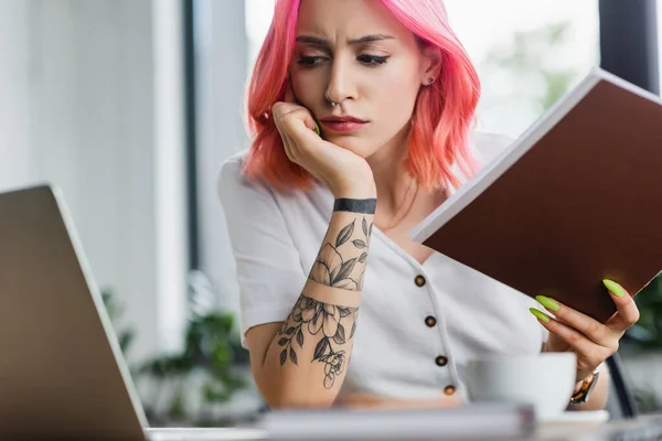 Unzufriedene Geschäftsfrau mit pinkfarbenen Haaren hält Ordner in der Hand und blickt auf Laptop — Stockfoto