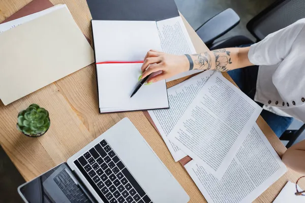 Vista recortada del gestor tatuado sosteniendo pluma cerca de documentos y computadora portátil en el escritorio - foto de stock