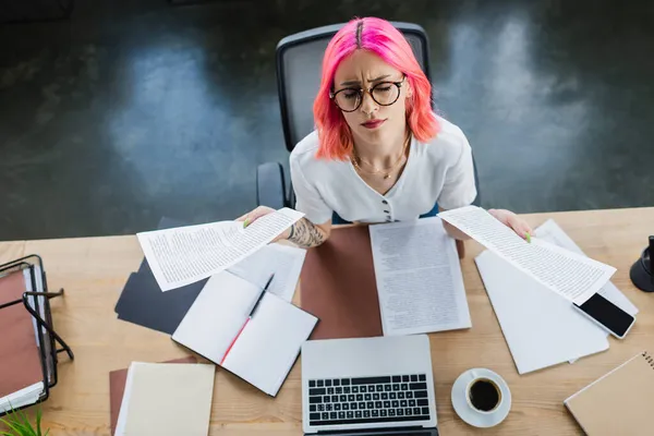 Vista superior de mujer de negocios cansada con documentos de celebración de pelo rosa cerca de gadgets en el escritorio - foto de stock