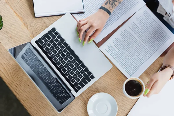 Cropped view of tattooed manager with green manicure holding cup of coffee near laptop and documents on desk — Stock Photo