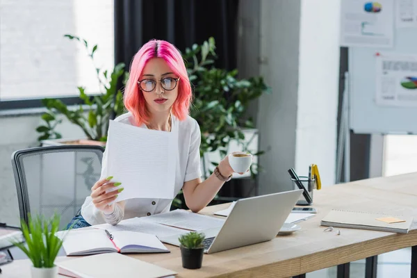 Gerente tatuado com cabelo rosa segurando xícara de café perto de laptop e documentos — Fotografia de Stock