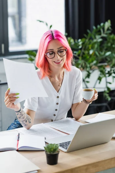 Gerente feliz com cabelo rosa segurando xícara de café perto de laptop e documentos — Fotografia de Stock