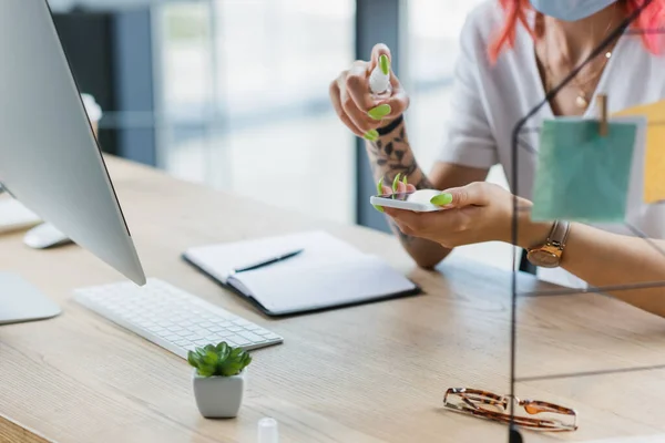 Ausgeschnittene Ansicht einer Frau in medizinischer Maske, die im Büro Desinfektionsmittel auf Smartphone sprüht — Stockfoto