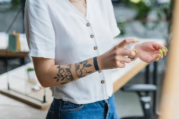 Vista cortada de empresária tatuada pulverizando desinfetante na mão no escritório — Fotografia de Stock