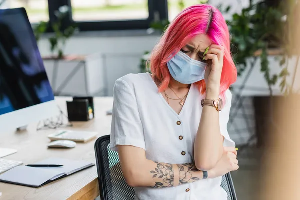 Mujer de negocios tatuada en máscara médica que tiene dolor de cabeza en la oficina - foto de stock