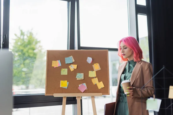 Junger Manager mit pinkfarbenen Haaren und durchdringendem Pappbecher in der Nähe von Karton mit klebrigen Notizen — Stockfoto