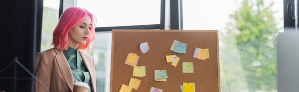 Young manager with pink hair and piercing holding paper cup near board with sticky notes, banner — Stock Photo