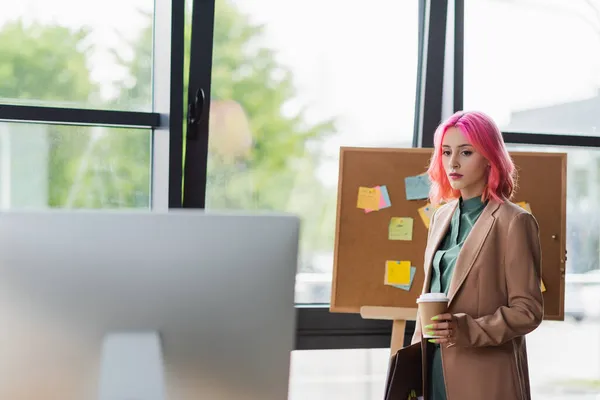 Jovem gerente com cabelo rosa e piercing segurando copo de papel e olhando para monitor de computador — Fotografia de Stock