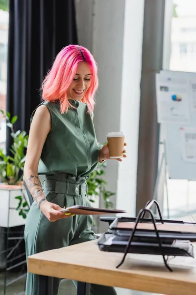 Cheerful manager with pink hair and piercing holding paper cup and folder near document tray — Stock Photo