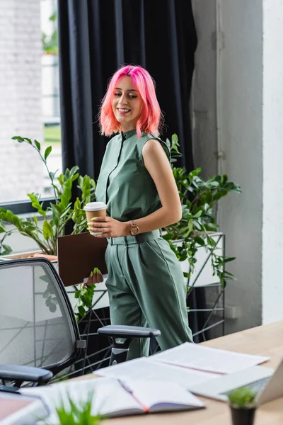 Fröhlicher Manager mit pinkfarbenen Haaren und Piercing, der Pappbecher und Ordner hält, während er auf den Schreibtisch blickt — Stockfoto