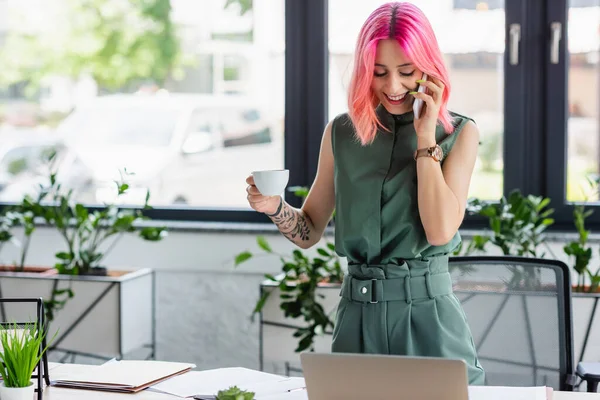 Веселий менеджер з рожевим волоссям і пірсингом тримає чашку кави під час розмови на смартфоні і дивлячись на ноутбук — стокове фото