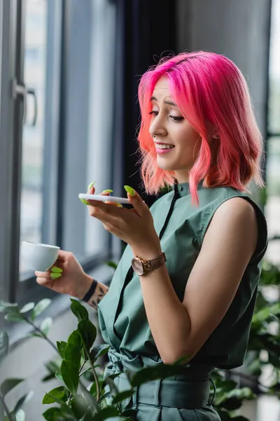 Femme d'affaires joyeuse avec les cheveux roses et perçant tenant tasse de café tout en enregistrant un message vocal sur smartphone — Photo de stock