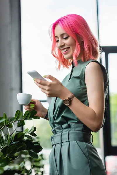 Alegre mujer de negocios con el pelo rosa y piercing taza de café mientras se utiliza el teléfono inteligente - foto de stock