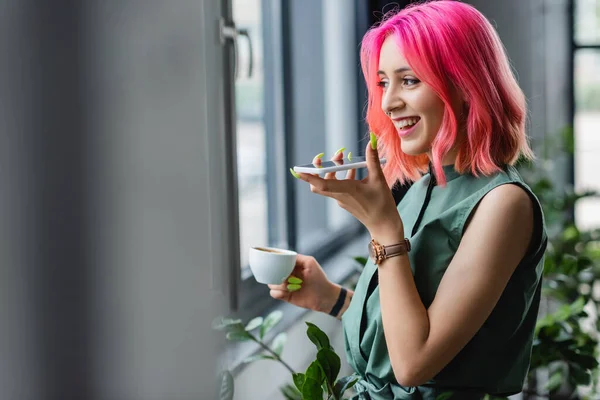 Радостная деловая женщина с розовыми волосами и пирсинг держа чашку кофе во время записи голосового сообщения на смартфон — стоковое фото