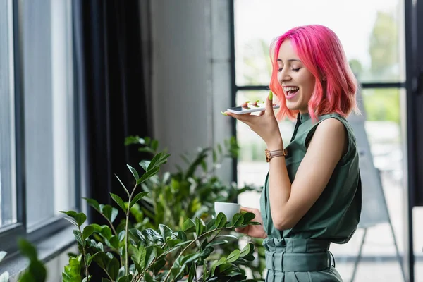 Positive businesswoman with pink hair and piercing holding cup of coffee while recording voice message on smartphone — Stock Photo