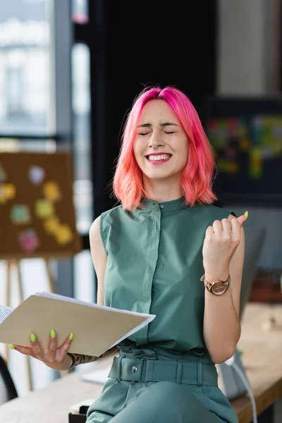 Alegre mujer de negocios con el pelo rosa y piercing carpeta de celebración en la oficina - foto de stock