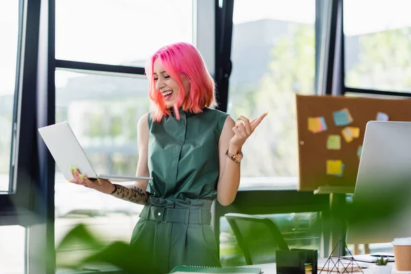 Femme d'affaires joyeuse avec les cheveux roses tenant ordinateur portable et pointant du doigt au bureau — Photo de stock