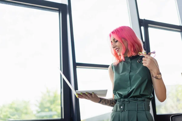 Cheerful businesswoman with pink hair holding laptop and eyeglasses in office — Stock Photo