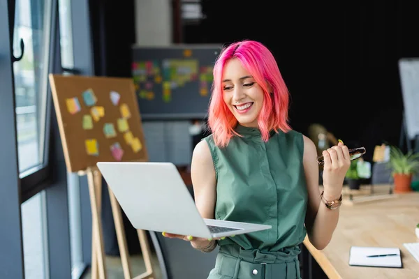 Радісна бізнес-леді з рожевим волоссям тримає ноутбук і окуляри в офісі — стокове фото