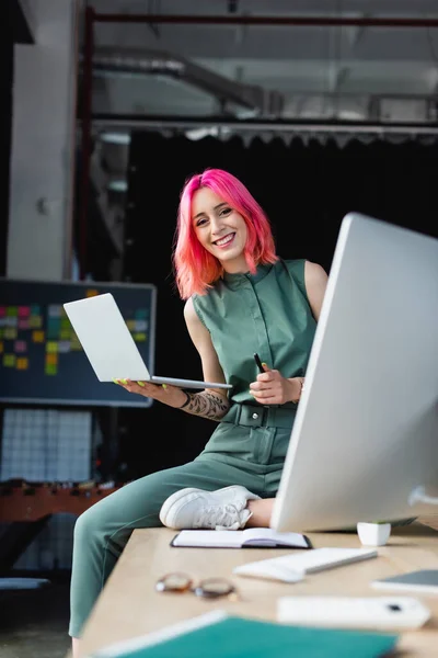Feliz mujer de negocios con el pelo rosa que sostiene la pluma y el ordenador portátil cerca del monitor de la computadora en la oficina - foto de stock