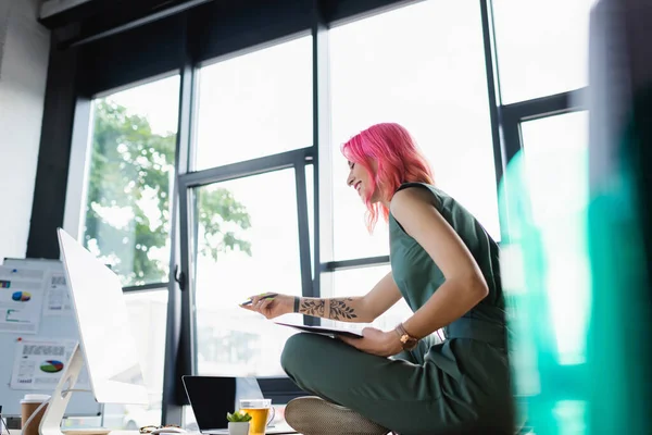 Femme d'affaires heureuse avec des cheveux roses tenant stylo tout en regardant moniteur d'ordinateur — Photo de stock