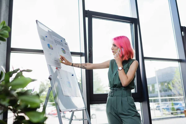 Geschäftsfrau mit pinkfarbenen Haaren telefoniert beim Blick auf Flipchart mit Diagrammen — Stockfoto