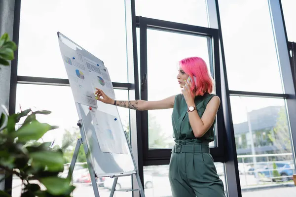 Donna d'affari con i capelli rosa che parla sul cellulare mentre guarda lavagna a fogli mobili con grafici — Foto stock