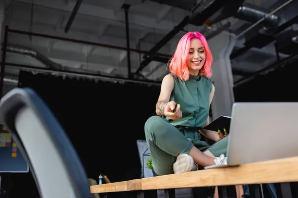 Happy businesswoman with pink hair holding pen and notebook while sitting on desk and looking at laptop — Stock Photo