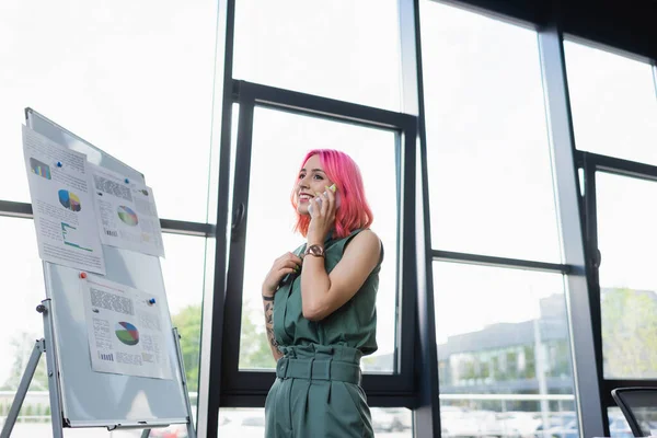 Felice donna d'affari con i capelli rosa parlando sul cellulare vicino lavagna a fogli mobili con grafici — Foto stock