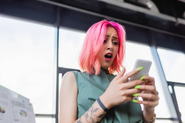 Vue à faible angle de la femme d'affaires choquée avec les cheveux roses en utilisant un smartphone — Photo de stock