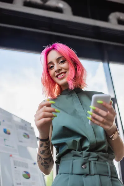 Low angle view of pleased businesswoman with pink hair holding smartphone — Stock Photo