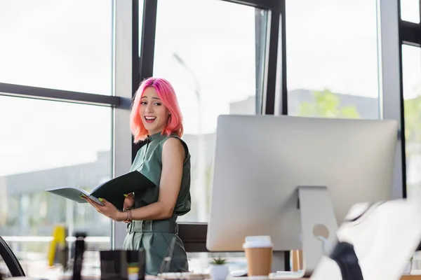Mujer de negocios sorprendida con la carpeta de retención de pelo rosa y mirando el monitor de la computadora - foto de stock