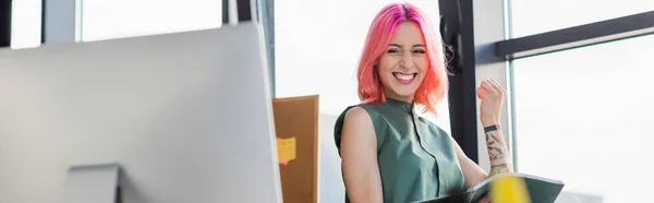 Cheerful businesswoman with pink hair holding folder and looking at computer monitor, banner — Stock Photo