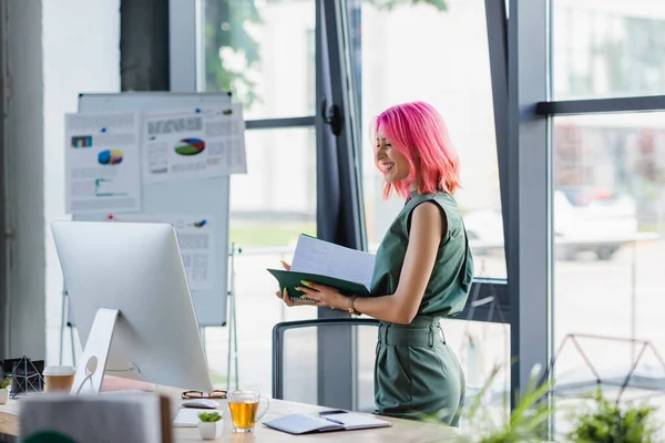 Alegre mujer de negocios con la carpeta de retención de pelo rosa y mirando el monitor de la computadora - foto de stock