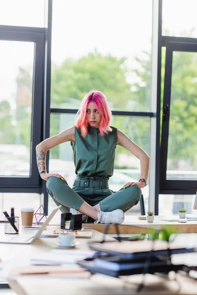 Displeased businesswoman with pink hair sitting with crossed legs on desk in office — Stock Photo