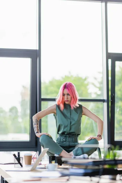 Деловая женщина с розовыми волосами сидит со скрещенными ногами на столе в офисе — стоковое фото
