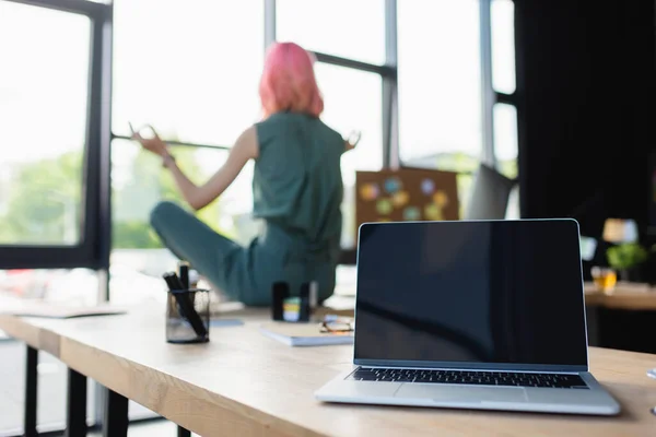 Ordinateur portable avec écran blanc près d'une femme d'affaires avec des cheveux roses méditant sur le bureau au bureau — Photo de stock