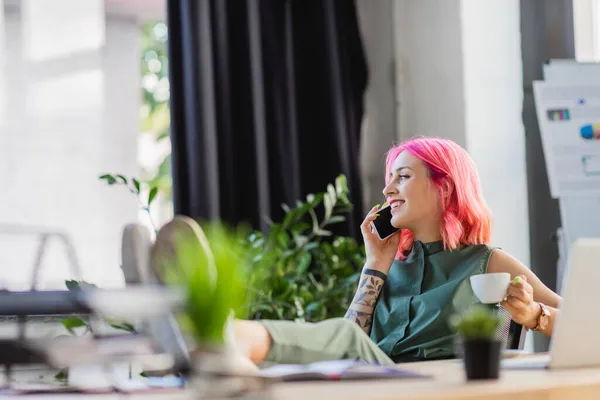Glückliche Geschäftsfrau mit rosafarbenen Haaren hält eine Tasse Kaffee in der Hand, während sie mit dem Smartphone spricht — Stockfoto