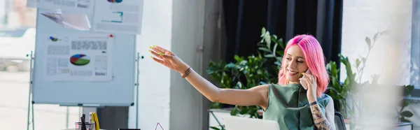 Alegre mujer de negocios con el pelo rosa hablando en el teléfono inteligente mientras lanza avión de papel, bandera - foto de stock